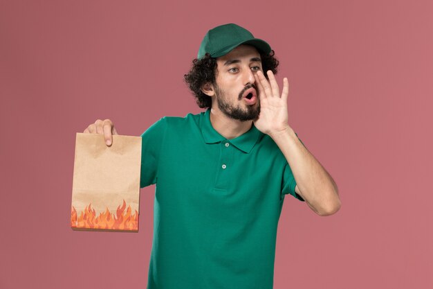 Front view male courier in green uniform and cape holding paper food package calling out on the pink background service worker uniform delivery job