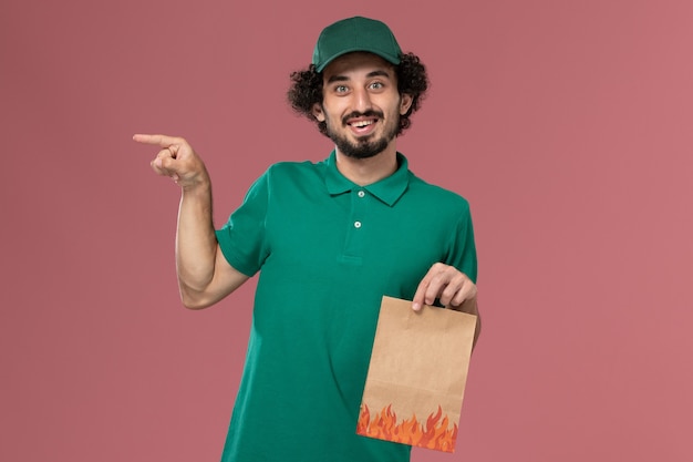 Вид спереди мужчина-курьер в зеленой форме и плаще, держащий бумажный пакет с едой на светло-розовом фоне, работа по доставке униформы работника службы