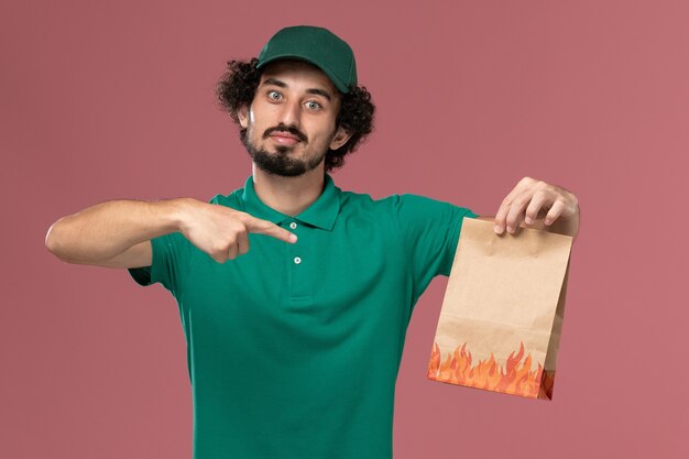 Front view male courier in green uniform and cape holding paper food package on pink background service uniform delivery job