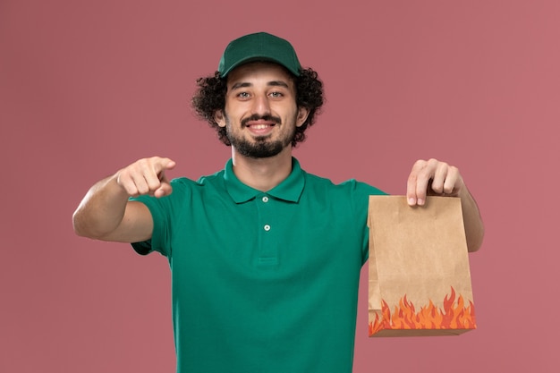 Front view male courier in green uniform and cape holding paper food package on the pink background service work uniform delivery job