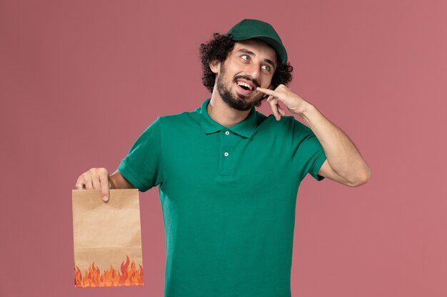 Front view male courier in green uniform and cape holding paper food package on the pink background service worker uniform delivery job work