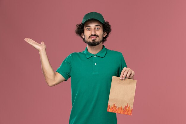 Front view male courier in green uniform and cape holding paper food package on the pink background service worker uniform delivery job