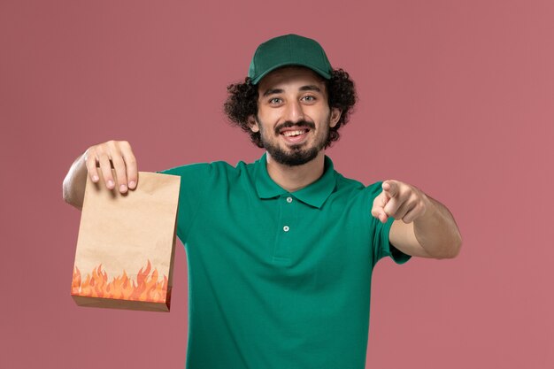 Front view male courier in green uniform and cape holding paper food package and smiling on the pink background service worker uniform delivery job