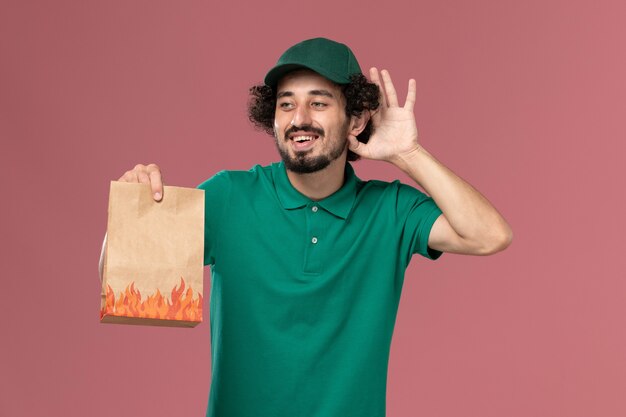 Вид спереди мужчина-курьер в зеленой форме и плаще, держащий бумажный пакет с едой, пытается услышать на розовом фоне работу по доставке униформы работника службы