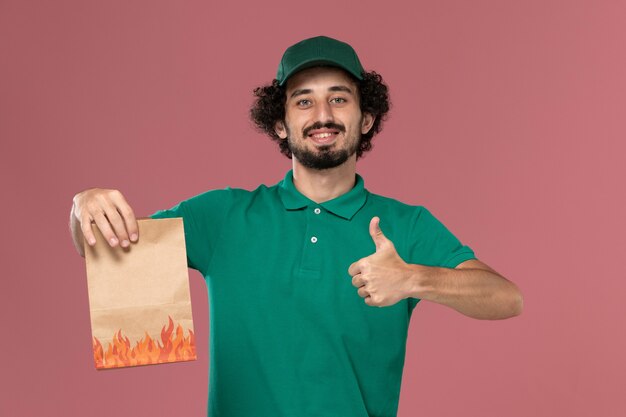 Front view male courier in green uniform and cape holding paper food package with smile on the pink background service worker uniform delivery job