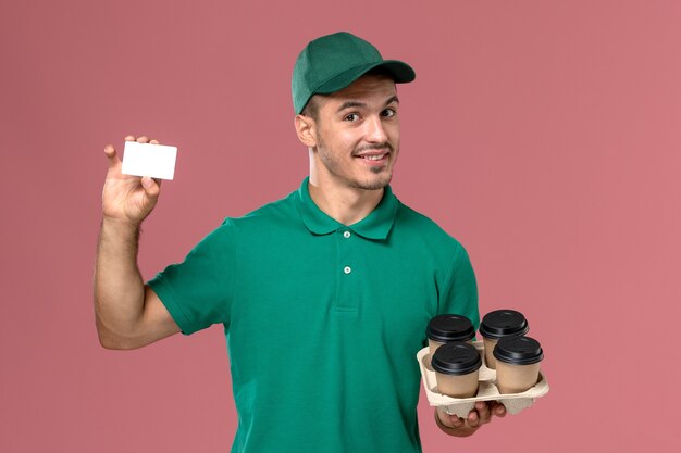 ピンクの背景に茶色のコーヒーカップとカードを保持している緑の制服の正面図男性宅配便