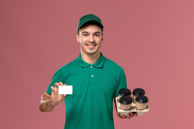 Front view male courier in green uniform holding brown coffee cups and card on pink background  