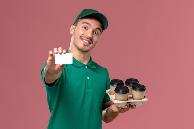 Front view male courier in green uniform holding brown coffee cups and card with smile on pink background  