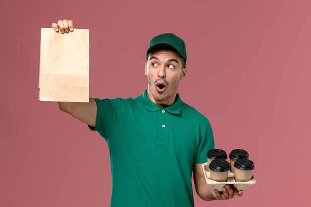 Front view male courier in green uniform holding brown coffee cups and food package on pink background