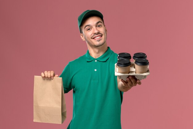 Front view male courier in green uniform holding brown coffee cups and food package on the pink background  
