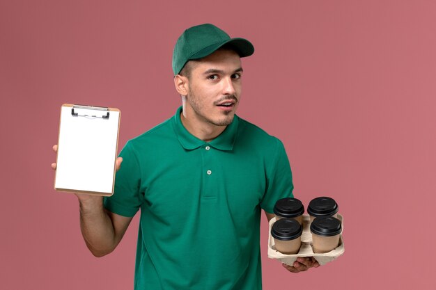 Front view male courier in green uniform holding brown coffee cups and notepad on pink background  