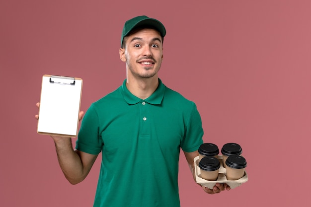 Foto gratuita corriere maschio di vista frontale in uniforme verde che tiene il blocco note marrone delle tazze di caffè sui precedenti rosa