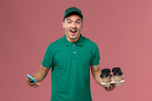 Front view male courier in green uniform holding brown coffee cups and phone on pink background  