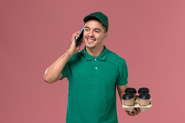 Front view male courier in green uniform holding brown coffee cups talking on the phone on pink desk 