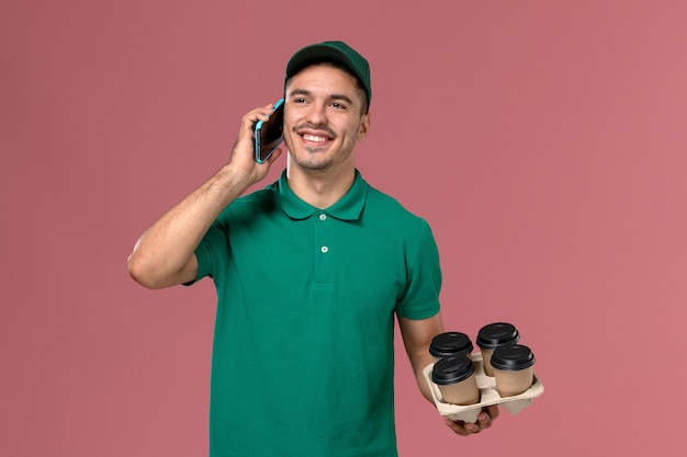 Front view male courier in green uniform holding brown coffee cups while talking on the phone on the pink desk