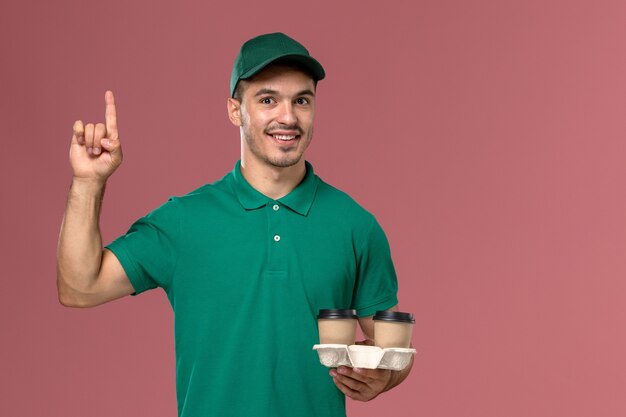 Front view male courier in green uniform holding brown delivery coffee cups on light-pink floor