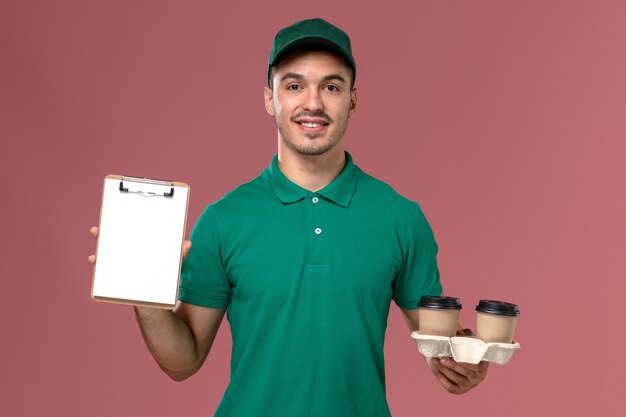 Курьер-мужчина, вид спереди в зеленой форме, держит коричневые кофейные чашки и блокнот на розовом столе