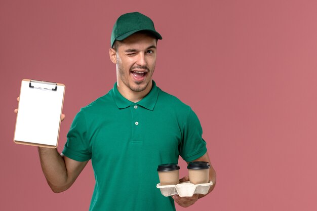 Курьер-мужчина в зеленой форме, вид спереди, держит коричневые кофейные чашки и блокнот, подмигивая на розовом столе