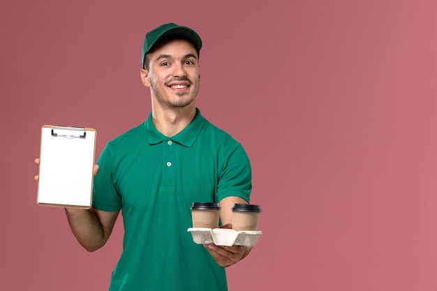 Курьер-мужчина в зеленой форме, вид спереди, держит коричневые кофейные чашки и блокнот с улыбкой на розовом столе