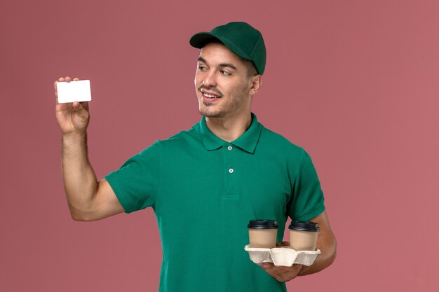 Front view male courier in green uniform holding brown delivery coffee cups and plastic card on the pink background