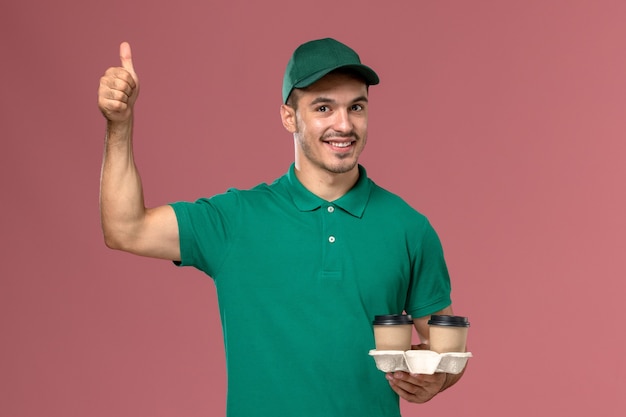 Front view male courier in green uniform holding brown delivery coffee cups smiling on light pink desk