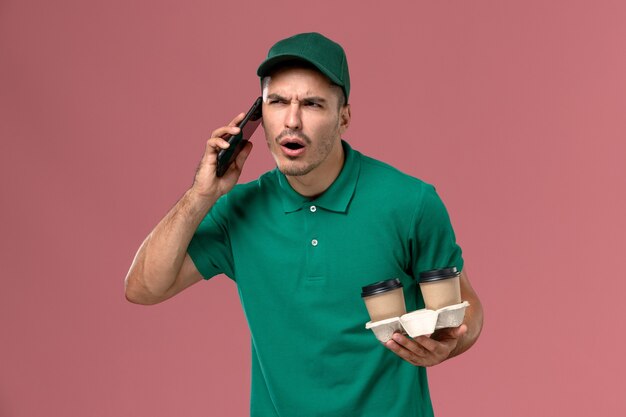 Front view male courier in green uniform holding brown delivery coffee cups and talking on the phone on pink background