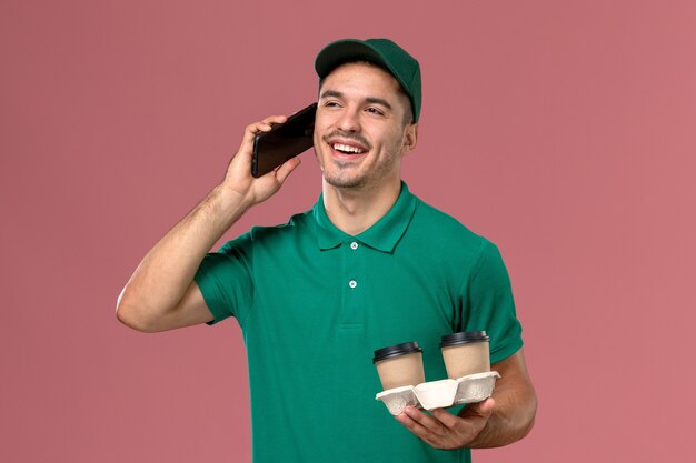 Front view male courier in green uniform holding brown delivery coffee cups and talking on the phone on pink desk