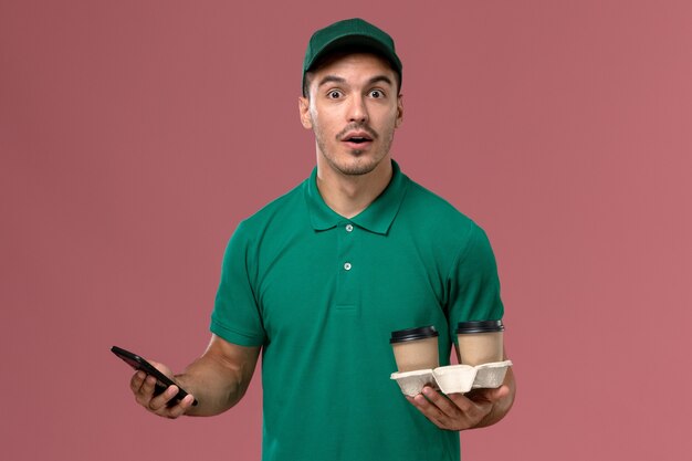 Курьер-мужчина в зеленой форме, вид спереди, держит коричневые кофейные чашки и использует телефон на розовом фоне