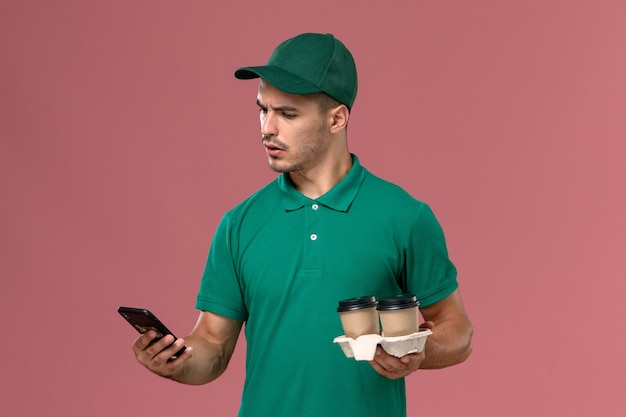 Курьер-мужчина в зеленой форме, вид спереди, держит коричневые кофейные чашки и разговаривает по телефону на розовом столе