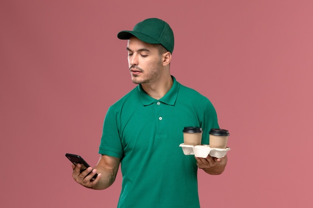 茶色の配達コーヒーカップを保持し、ピンクの机の上で電話を使用して緑の制服を着た正面図の男性宅配便