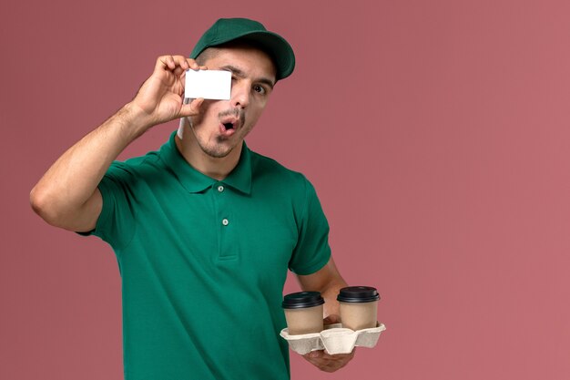 Front view male courier in green uniform holding brown delivery coffee cups and white plastic card on pink desk