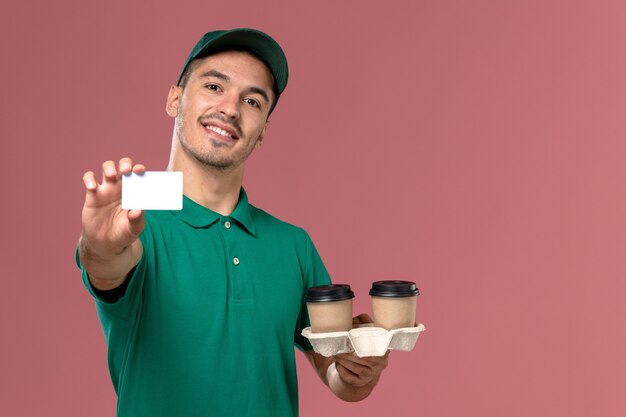 Front view male courier in green uniform holding brown delivery coffee cups and white plastic card smiling on pink desk