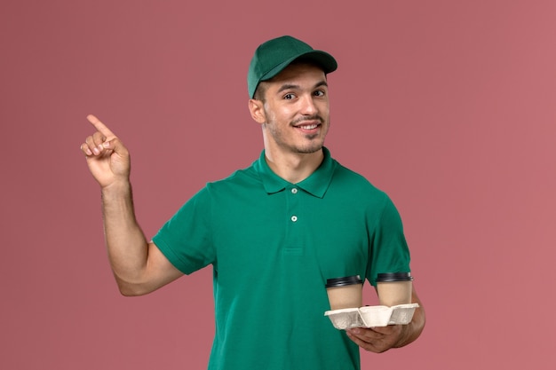 Курьер-мужчина в зеленой форме с улыбкой держит коричневые кофейные чашки на розовом столе