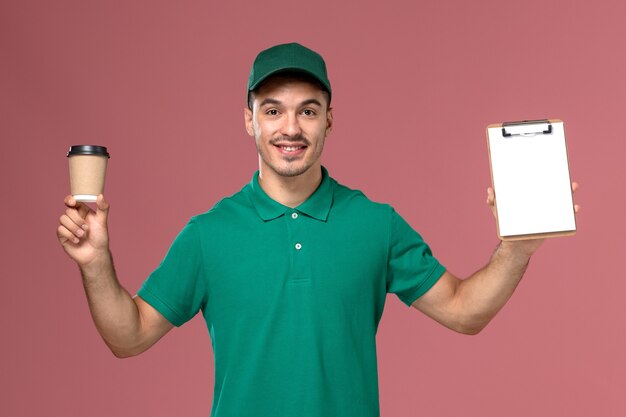 Front view male courier in green uniform holding coffee cup and notepad on pink background