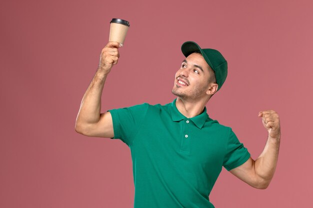 Front view male courier in green uniform holding delivery coffee cup and flexing on the pink desk