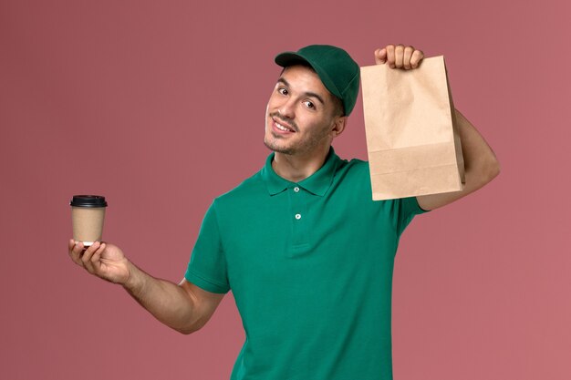 Front view male courier in green uniform holding delivery coffee cup and food package on the light-pink background