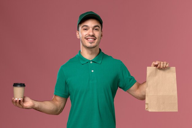 Front view male courier in green uniform holding delivery coffee cup and food package on light-pink desk