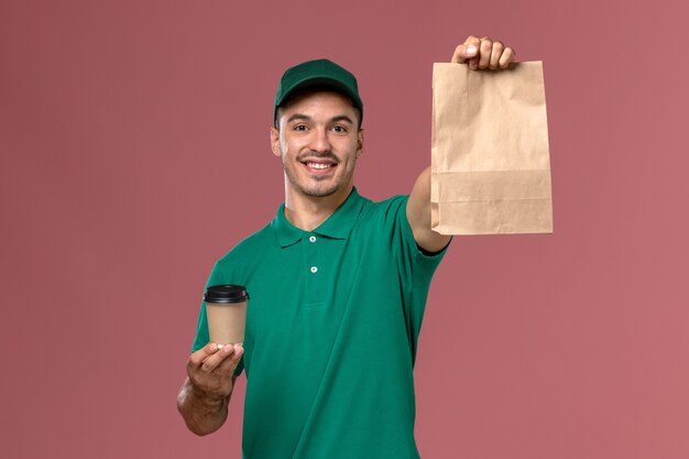 Front view male courier in green uniform holding delivery coffee cup and food package with smile on light-pink background