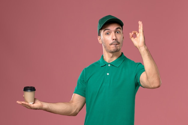 Front view male courier in green uniform holding delivery coffee cup on light pink background 