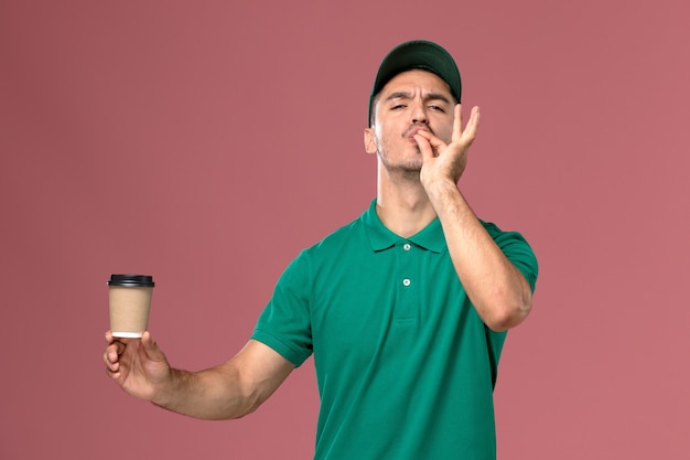 Курьер-мужчина в зеленой форме, вид спереди, держит чашку кофе на светло-розовом столе