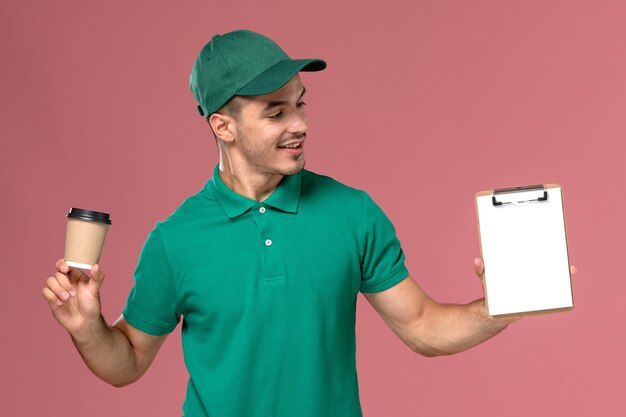 Front view male courier in green uniform holding delivery coffee cup and notepad on the pink desk  