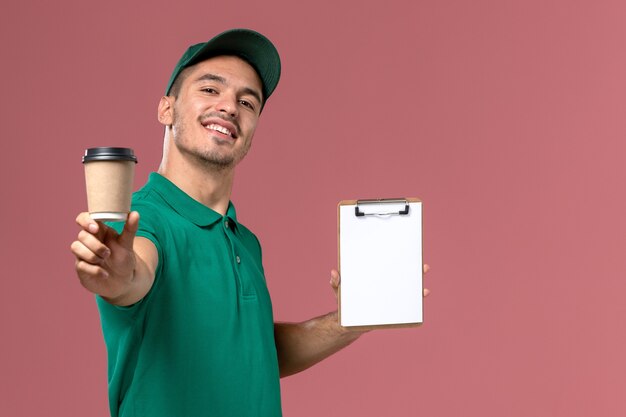 Front view male courier in green uniform holding delivery coffee cup and notepad smiling on the light-pink desk  