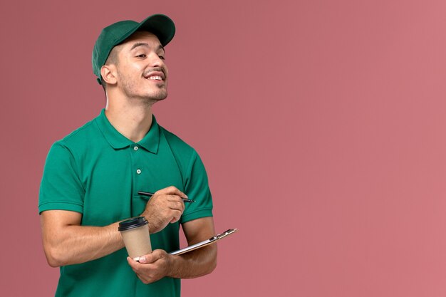 Курьер-мужчина в зеленой униформе, вид спереди, держит чашку кофе и пишет блокнот на светло-розовом столе