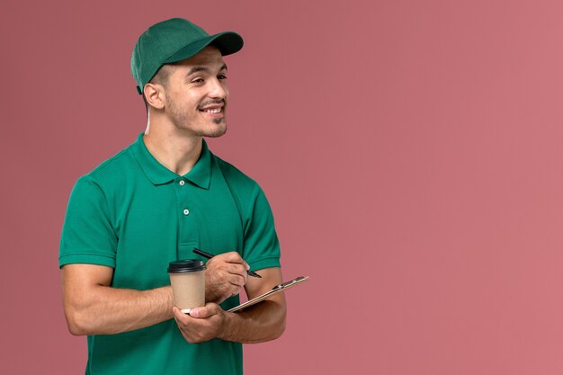 淡いピンクの机の上に配達コーヒーカップとメモ帳を保持している緑の制服を着た正面図の男性宅配便