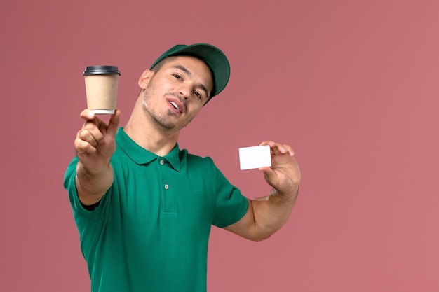Курьер-мужчина в зеленой форме, вид спереди, держит чашку кофе с белой карточкой на розовом столе