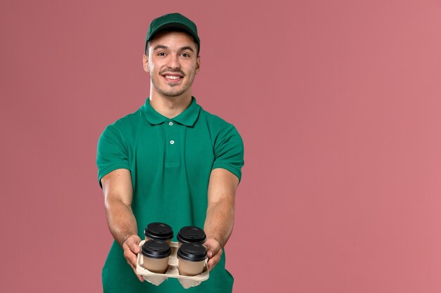 ピンクの背景にそれらを配信する配信コーヒーカップを保持している緑の制服を着た正面図男性宅配便