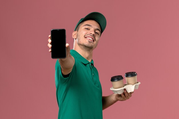 Front view male courier in green uniform holding delivery coffee cups and phone on pink background 
