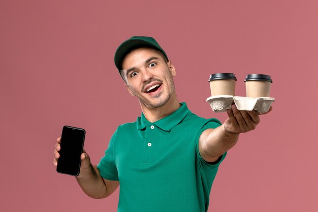 Front view male courier in green uniform holding delivery coffee cups and phone on the pink background 