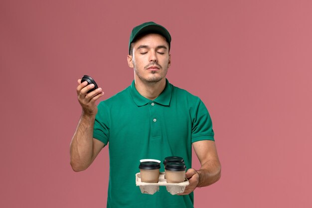 Front view male courier in green uniform holding delivery coffee cups smelling their scent on pink desk 