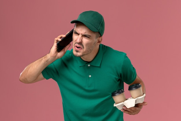 Front view male courier in green uniform holding delivery coffee cups and talking on the phone on pink background 